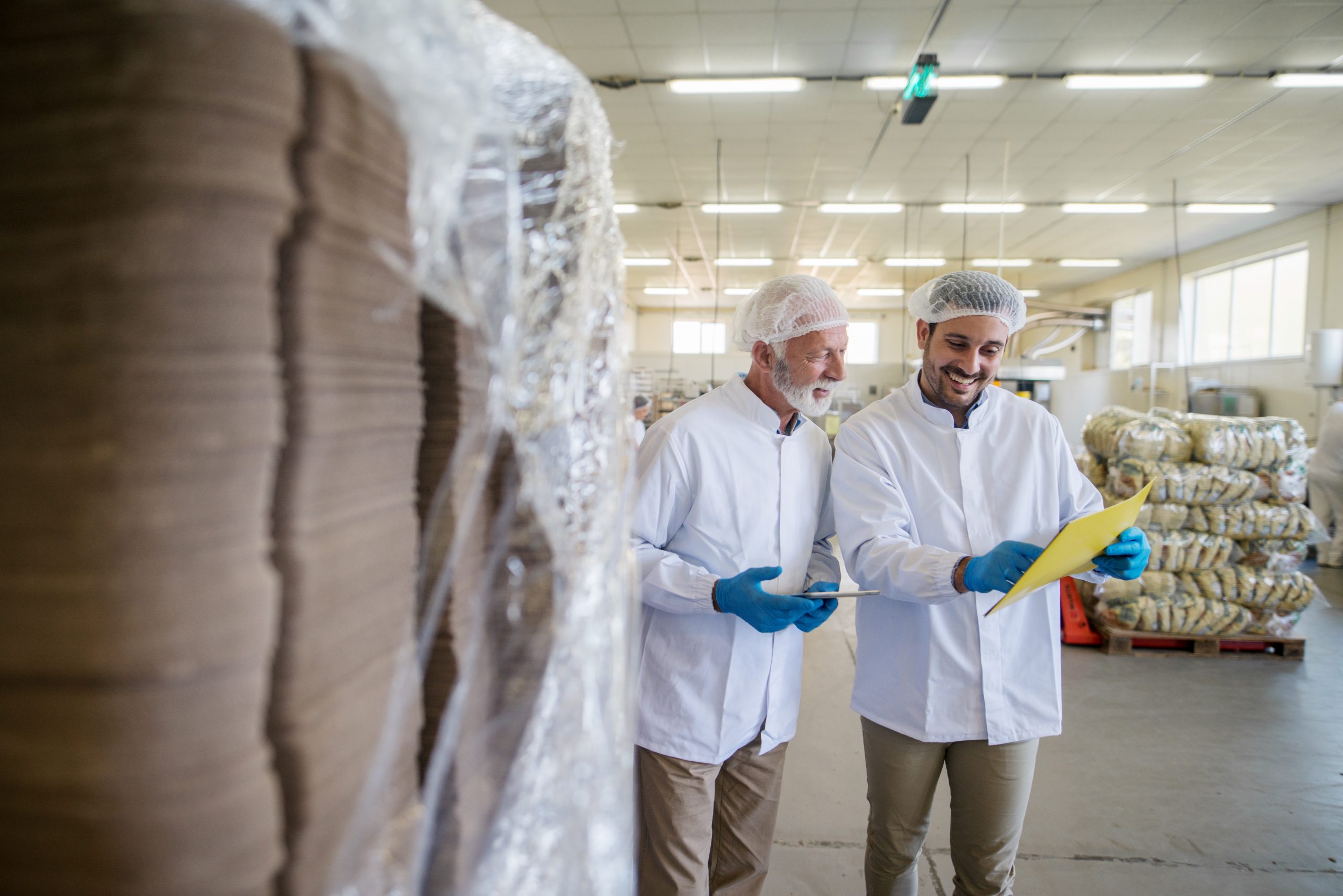 Two male managers in sterile clothes standing in warehouse and checking documents of package prepared for transport.