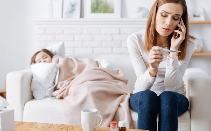 We have troubles. Troubled caring mother calling a doctor while her sick daughter lyign on the couch in the background