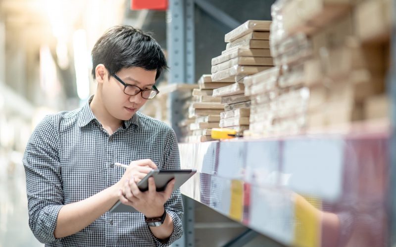 Young Asian man worker doing stocktaking of product in cardboard box on shelves in warehouse by using digital tablet and pen. Physical inventory count concept
