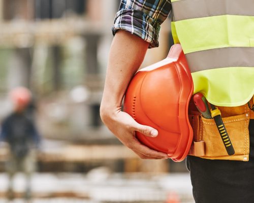 Construction worker. Cropped photo of male professional builder in working uniform with construction tools holding a safety red helmet while standing outdoor of construction site. Industry concept. Building concept. Technology concept.