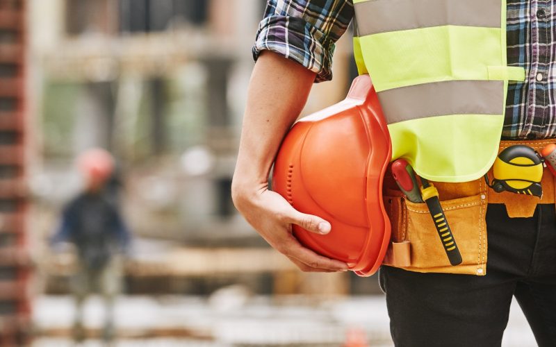 Construction worker. Cropped photo of male professional builder in working uniform with construction tools holding a safety red helmet while standing outdoor of construction site. Industry concept. Building concept. Technology concept.