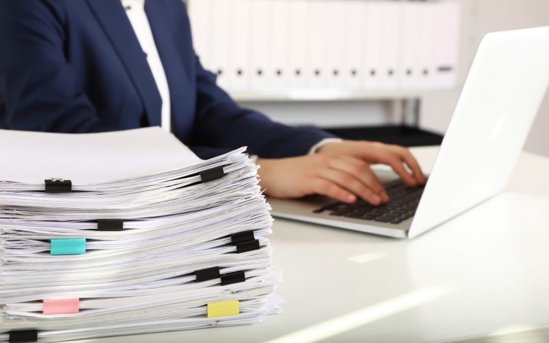 Stack of documents and woman working with laptop at table in office, closeup