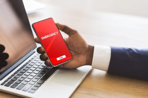 Safety Concept. Male hand holding smart phone with emergency number on red screen over office background and laptop