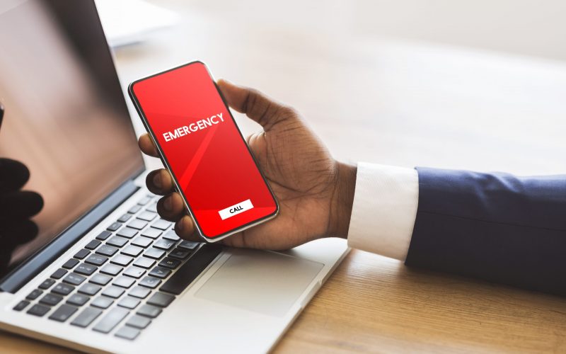 Safety Concept. Male hand holding smart phone with emergency number on red screen over office background and laptop