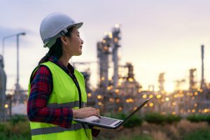 Asian woman petrochemical engineer working at night with laptop Inside oil and gas refinery plant industry factory at night for inspector safety quality control.
