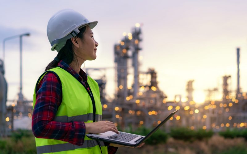 Asian woman petrochemical engineer working at night with laptop Inside oil and gas refinery plant industry factory at night for inspector safety quality control.