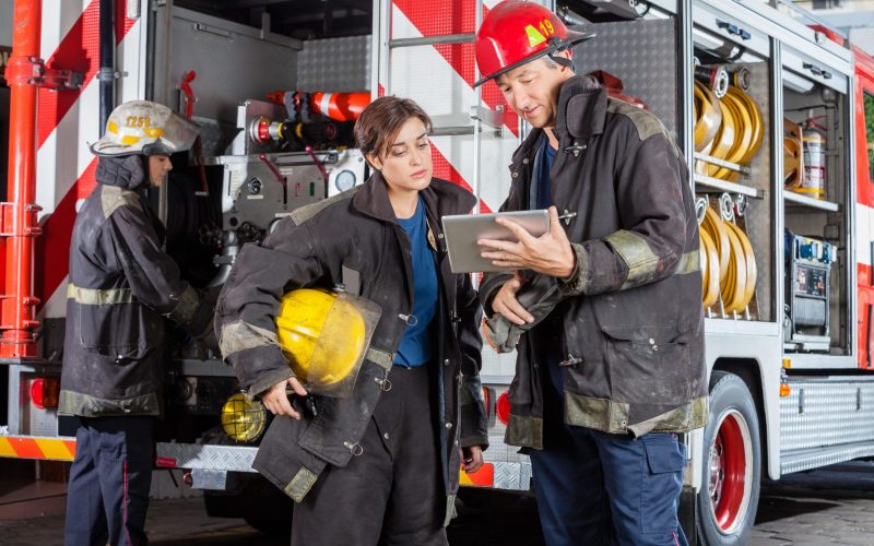 Young and mature firefighters using tablet computer against truck at fire station